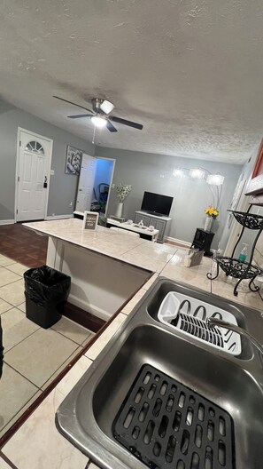 A kitchen with a built-in dishwasher, a sink with a dish rack, a ceiling fan, and a flat-screen TV. - Cozy 1-bedroom apartment in beautiful Snellville Town (Snellville)