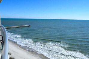 Ein Strand mit einem hölzernen Steg, der sich ins Meer erstreckt, einem klaren, blauen Himmel und sanften Wellen am Ufer.