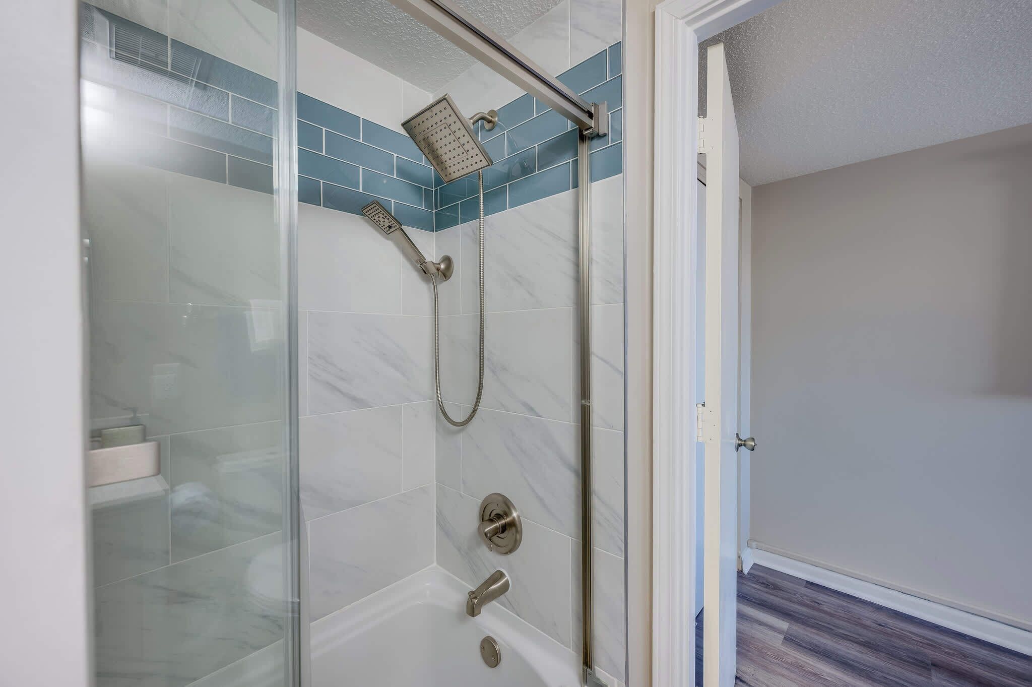 A glass-enclosed shower with a handheld showerhead and a fixed showerhead, a glass door, and a tiled floor.