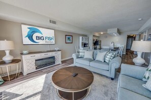 A modern living room with a light blue sofa, a round coffee table, and a TV mounted on the wall displaying 'BIG FISH Rentals'. - The Oceans 2 BR Condo Oceanfront Views Large Condo (North Myrtle Beach)