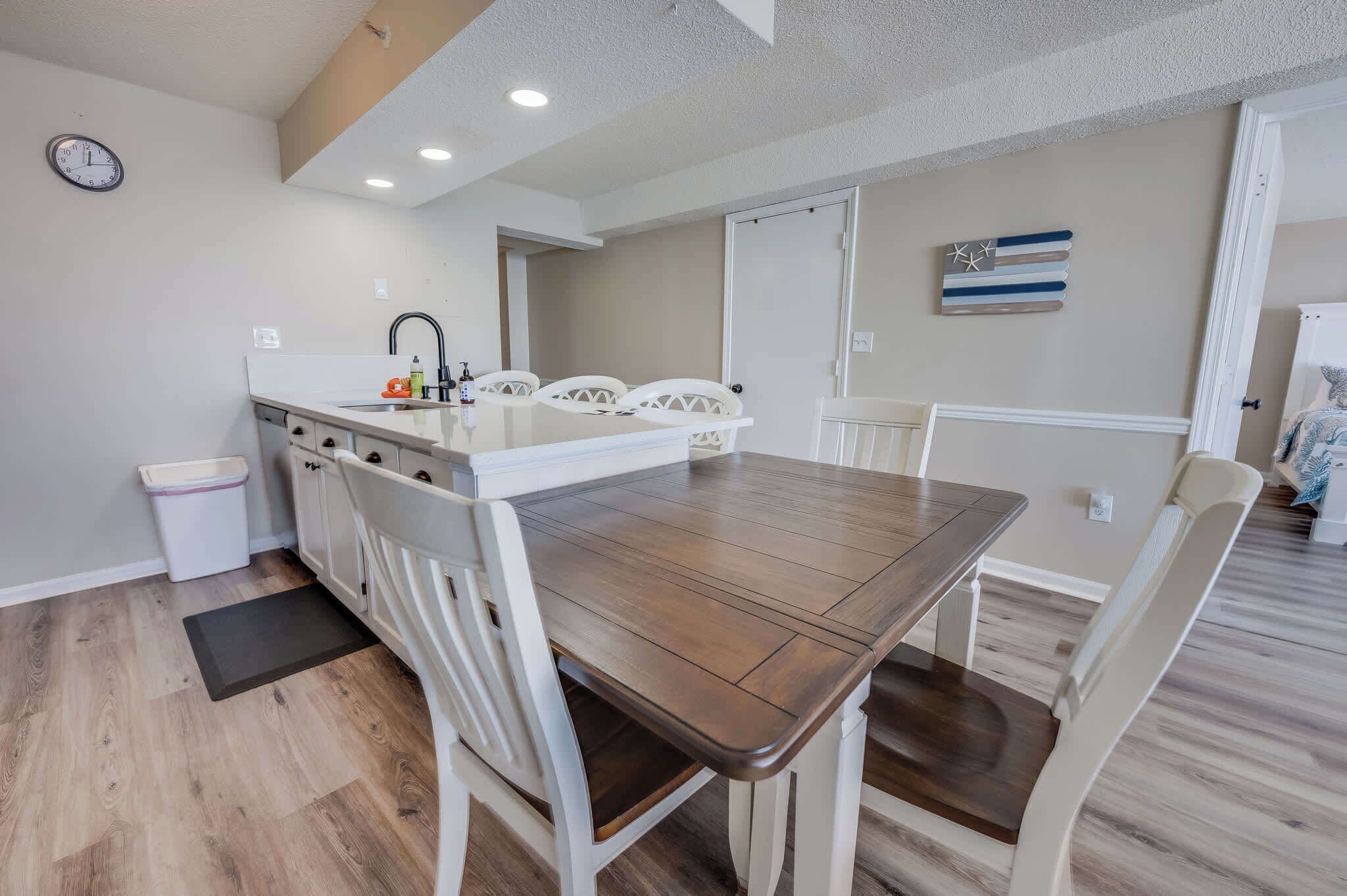 A dining area with a wooden table and white chairs, a kitchen with a sink and cabinets, and a clock on the wall.