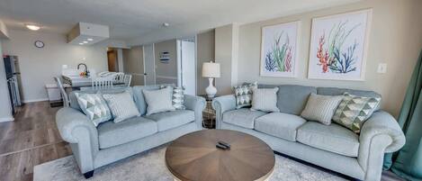 A modern living room with two light grey sofas, a round coffee table, and a kitchen area in the background.