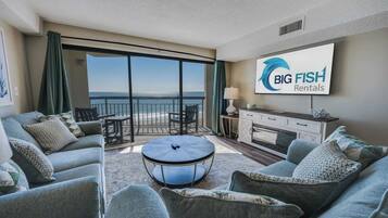 Un salón moderno con un amplio ventanal corredero que da a un balcón con vista al océano. Hay una mesa de centro redonda, un sofá y un televisor montado en la pared mostrando el logotipo de Big Fish Rentals.