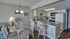 A compact kitchen with white cabinets, a central island, and a dining area with wicker chairs. - Direct Oceanfront 1BR, Pools at Sands Ocean Annies (Myrtle Beach)