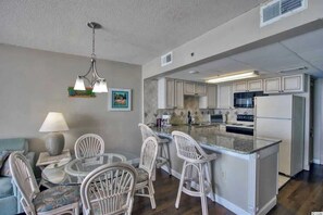 A compact kitchen with white cabinets, a central island, and a dining area with wicker chairs. - Direct Oceanfront 1BR, Pools at Sands Ocean Annies (Myrtle Beach)