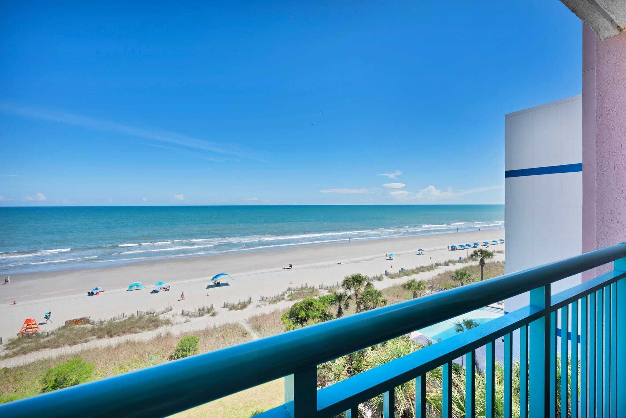 A balcony with a view of a beach, a pool, and a building with a blue stripe.