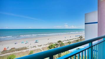 A balcony with a view of a beach, a pool, and a building with a blue stripe.