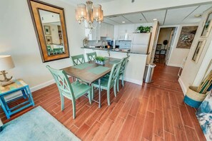 Un coin repas avec une table en bois et des chaises vertes, un lustre et une cuisine en arrière-plan.