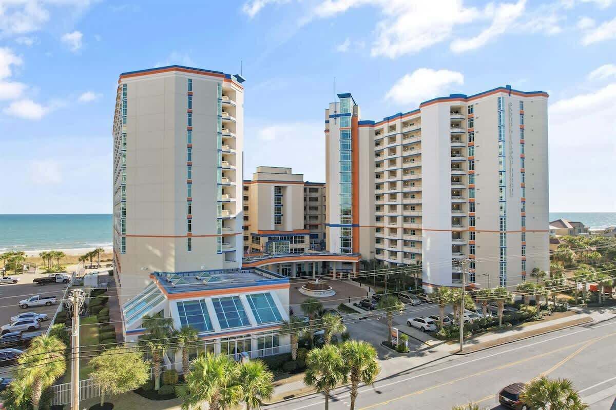 A multi-story hotel with a central glass atrium, adjacent to a beach.