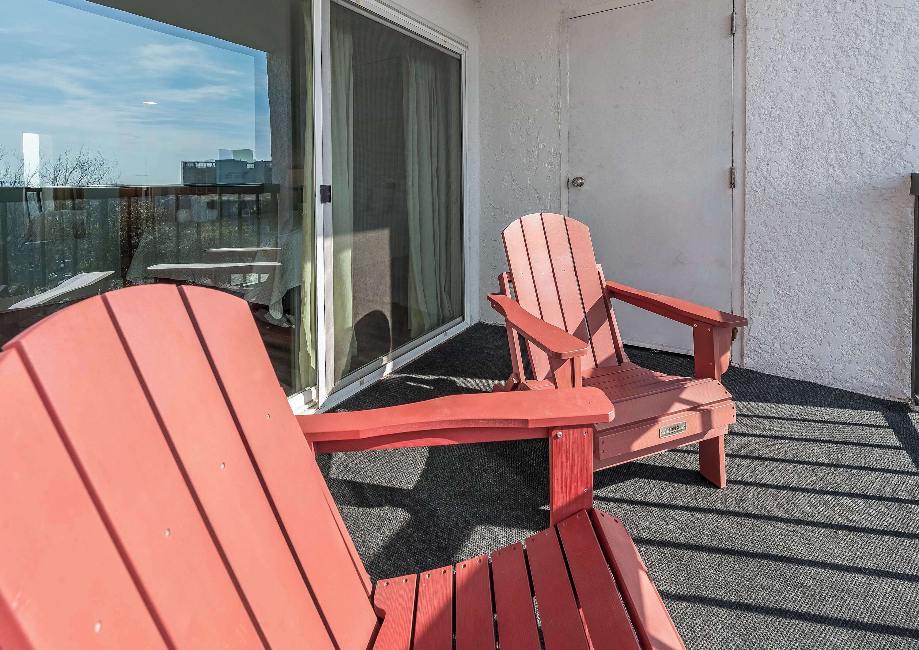 A balcony with two red Adirondack chairs, a sliding glass door, and a view of the cityscape.