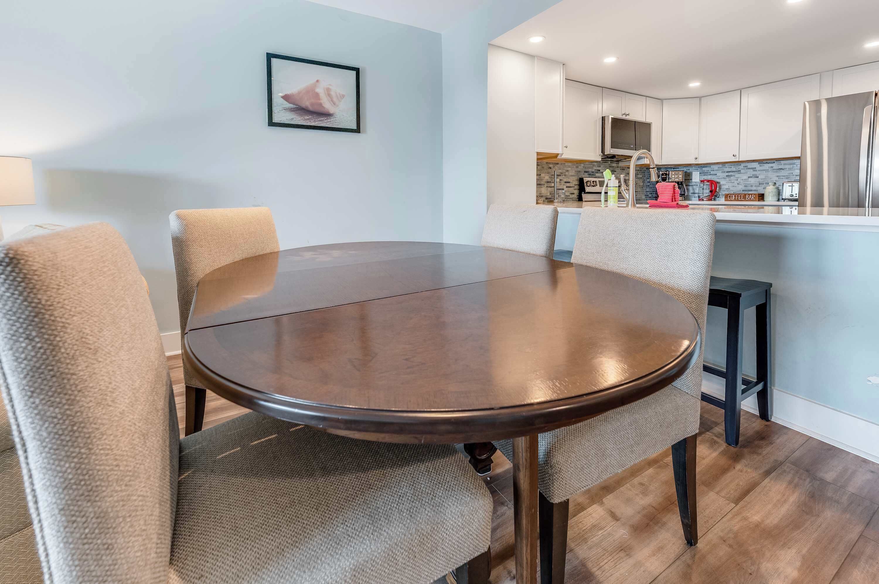 Un comedor moderno con una mesa redonda y cuatro sillas, una cocina con armarios blancos y un refrigerador de acero inoxidable, y un cuadro en la pared.