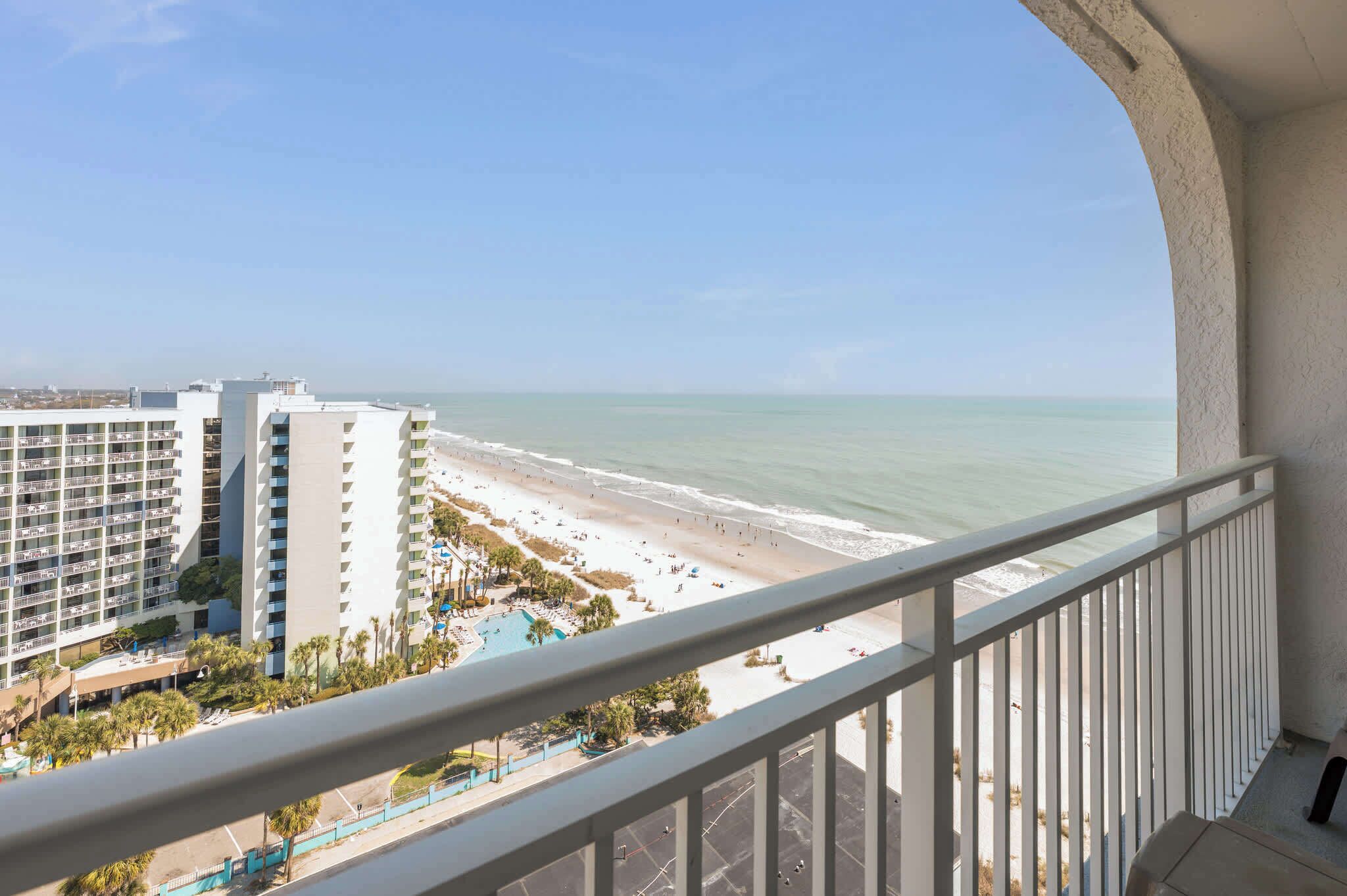 A balcony with a view of a beach, a pool, and a hotel building.