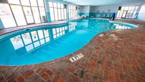 Una piscina interna con acqua cristallina e azzurra, circondata da pavimento in mattoni e ampie finestre.