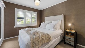 A bedroom with a large bed, a window with blinds, a bedside lamp, and a small wooden nightstand. - The One - Kinloch (Kinloch)
