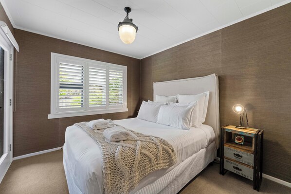 Une chambre avec un grand lit, une fenêtre avec des stores, une lampe de chevet et une petite table de nuit en bois.