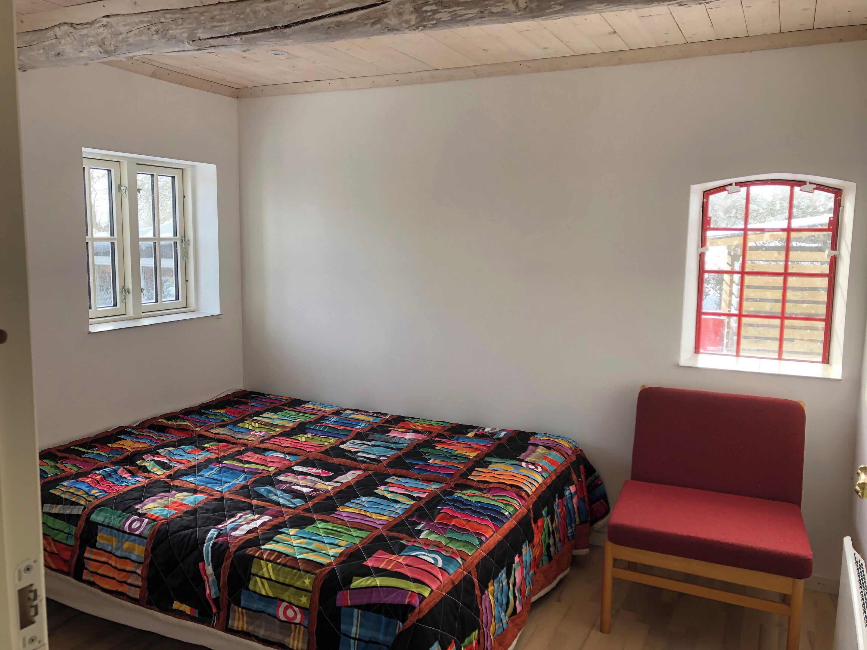 Une chambre à coucher avec un lit, une chaise rouge, deux fenêtres et un plafond en bois.