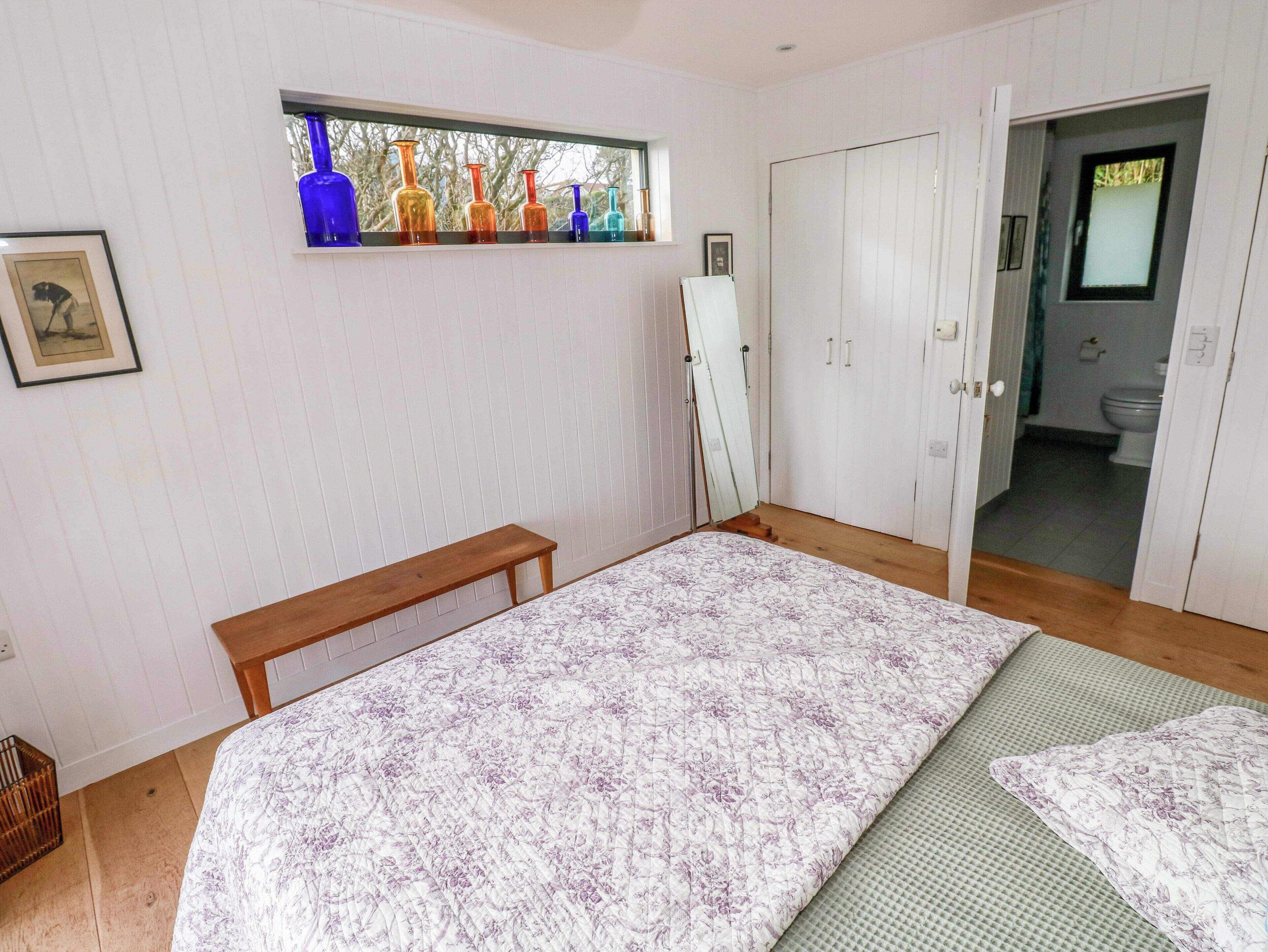 A bedroom with a bed, a wooden bench, a window with colorful vases, and a door leading to a bathroom.