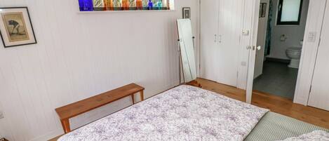 A bedroom with a bed, a wooden bench, a window with colorful vases, and a door leading to a bathroom.