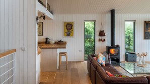 A modern living room with a glass coffee table, a brown leather sofa, a wooden dining table, and a black wood-burning stove. - Green Shutters (Pembroke)