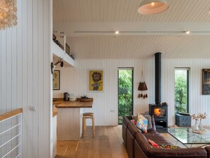 A modern living room with a glass coffee table, a brown leather sofa, a wooden dining table, and a black wood-burning stove. - Green Shutters (Pembroke)