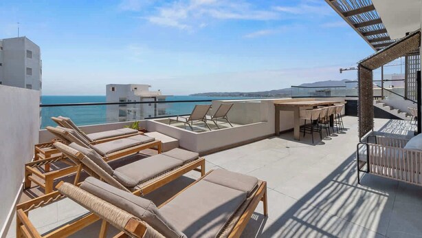 Terraza en la azotea con sillones de sol, una mesa y sillas, con vistas a un cuerpo de agua y colinas distantes.