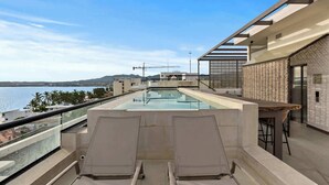 Zona de piscina en la azotea con dos sillones, un bar y vistas al mar.