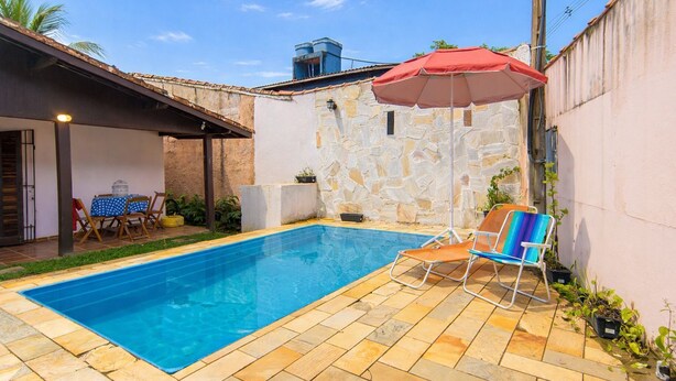 A rectangular swimming pool with tiled flooring, a stone wall, and a red umbrella. There are two lounge chairs and a table with chairs. - Casa com Piscina e rea Gourmet em Boi Ucanga (São Sebastião)