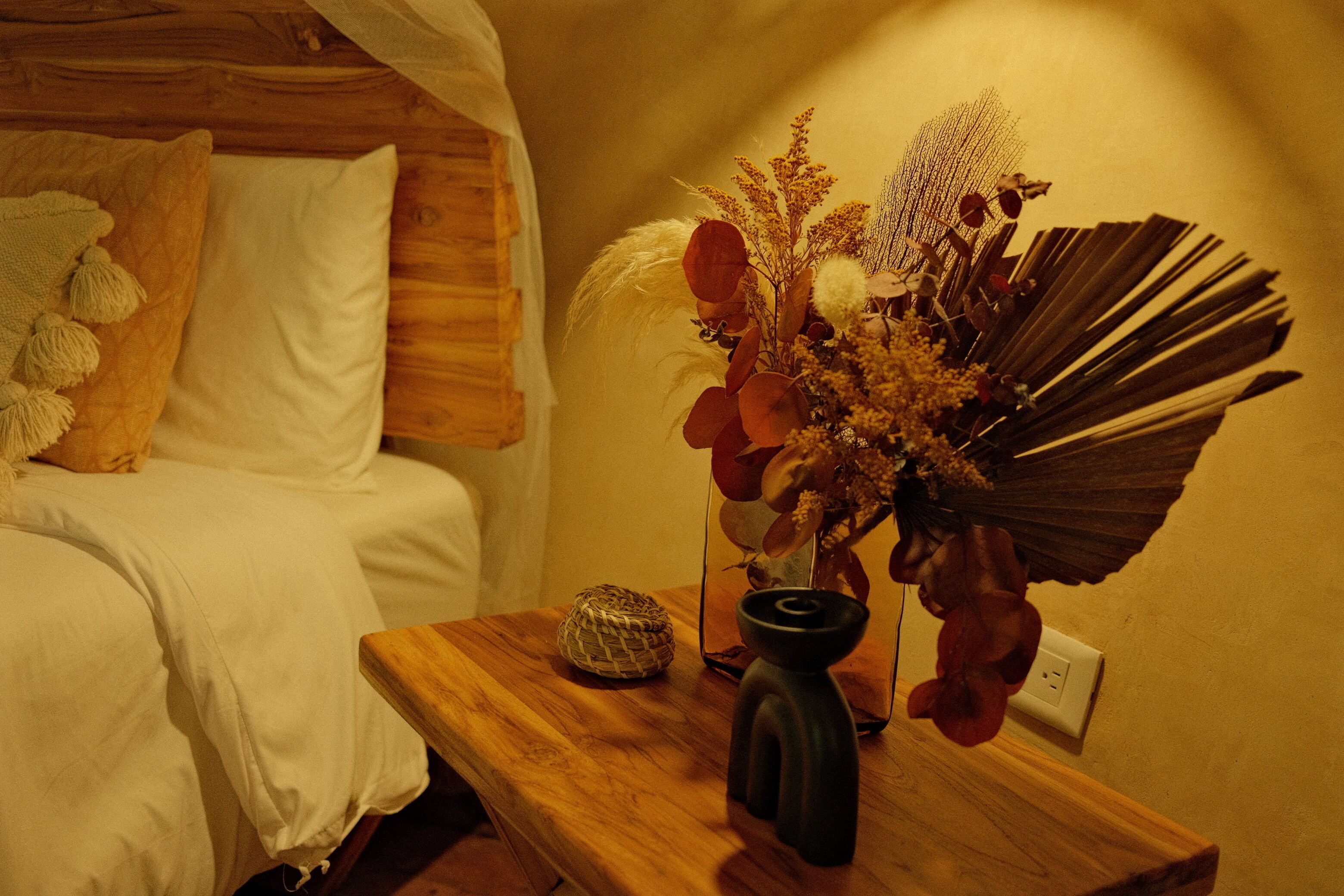 A bedroom with a wooden headboard, a bedside table with a vase of dried flowers, and a woven basket.
