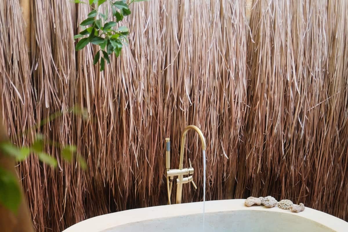 A freestanding bathtub with a faucet, surrounded by pebbles, against a backdrop of a straw-like wall.
