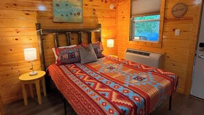 Um quarto acolhedor com ventoinha de madeira no teto, uma cama com manta estampada, duas luminárias de cabeceira e uma janela com vista para árvores.
