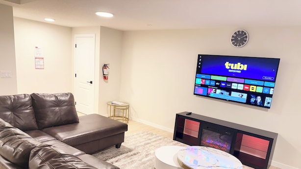 A living room with a sectional sofa, a flat-screen TV mounted on the wall, a small round table, and a fireplace unit. - The Luxe Haven - Stylish 2BR | 2BA Retreat •Comfort • Privacy • Perfect Location (Calgary)
