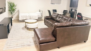 Una sala moderna con un sofá de cuero marrón, una mesita redonda, una cama con ropa de cama blanca, una mesa de comedor con sillas y una planta en maceta.