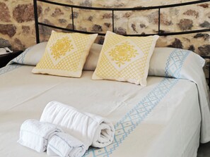 A neatly made bed with patterned pillows and a rolled towel on a bed with a stone wall background. - Locanda Jannas (Orgosolo)