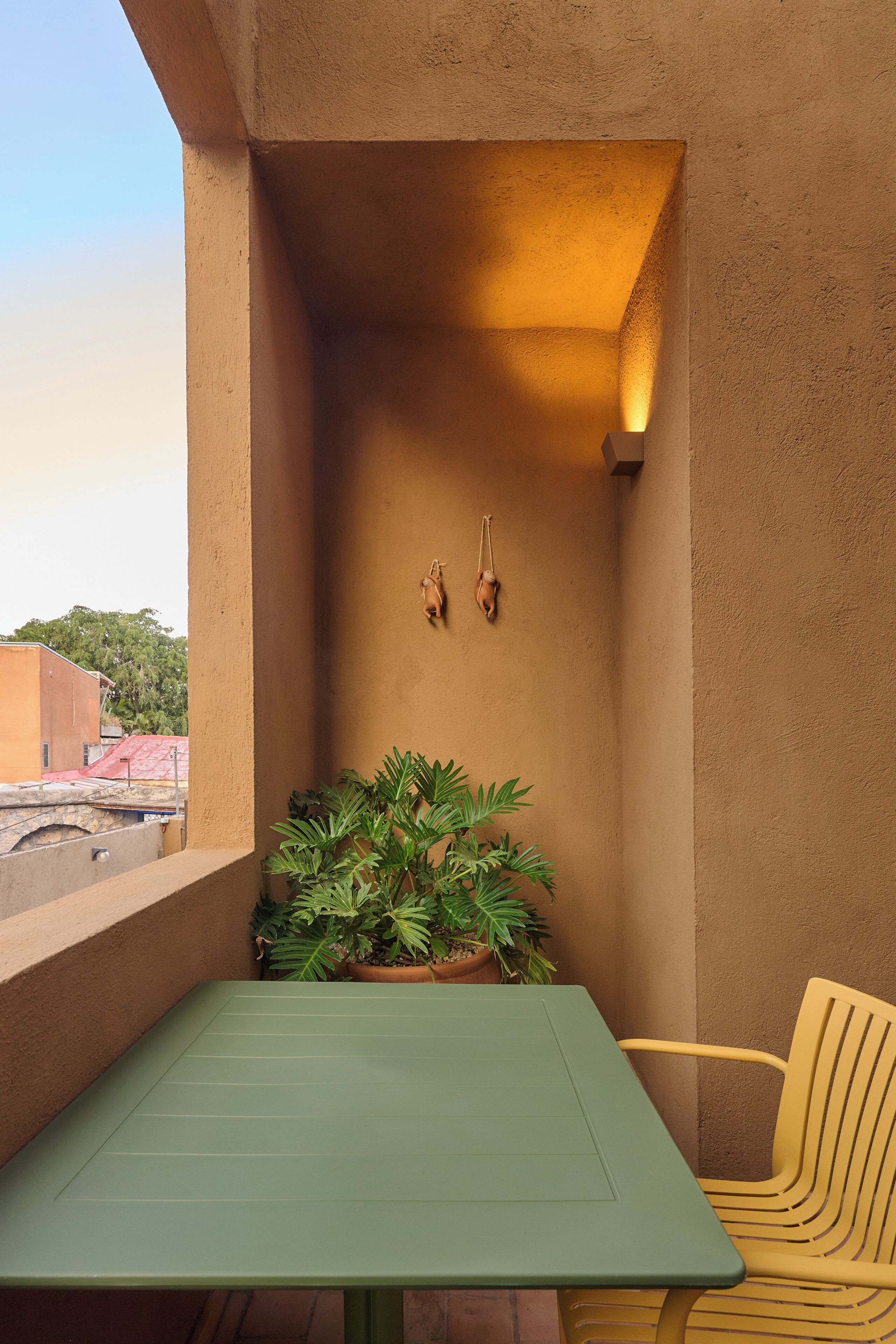 A balcony with a green table, a yellow chair, a potted plant, and a wall-mounted light fixture.