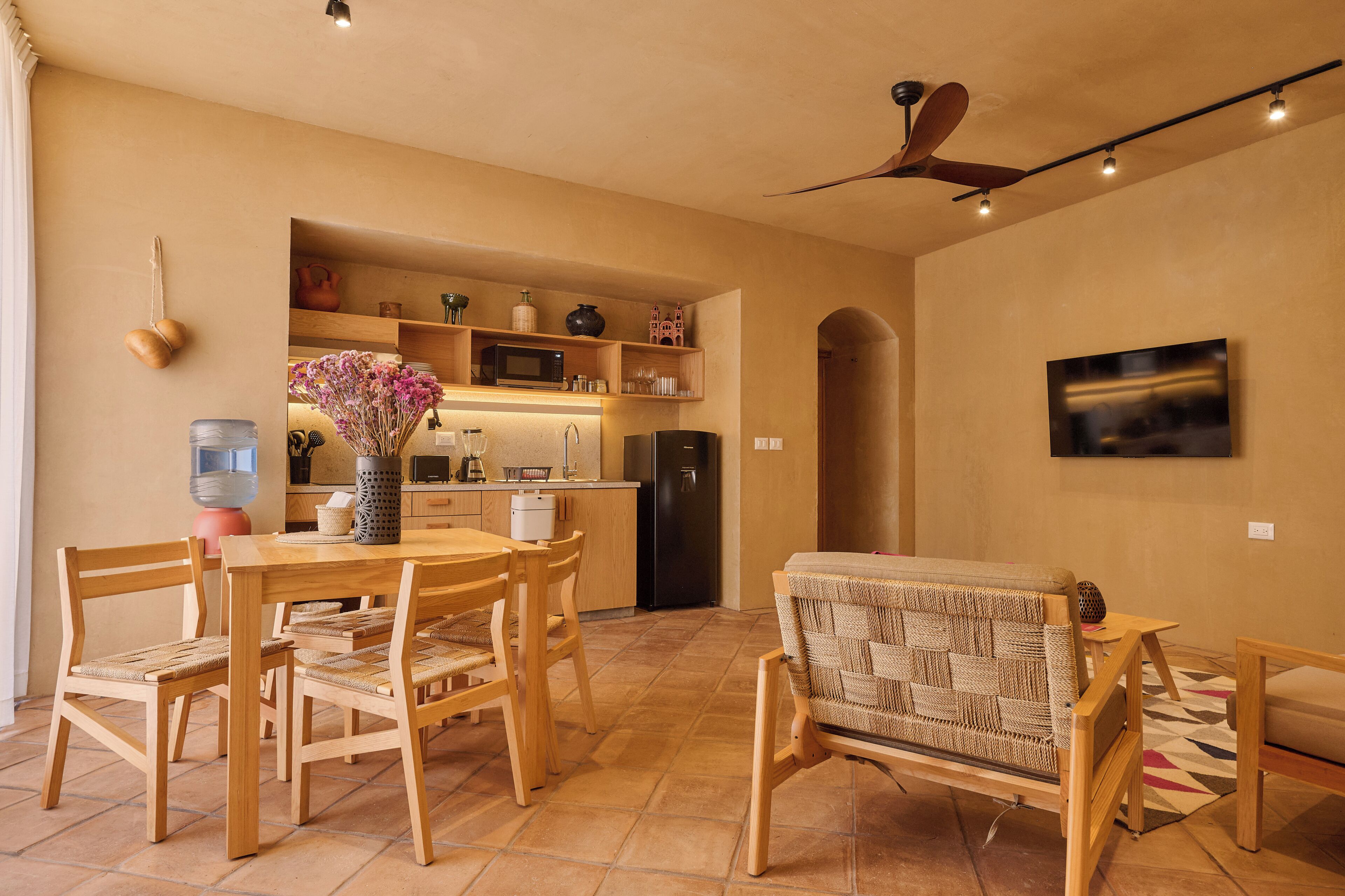 A modern kitchen with wooden cabinets, a microwave, and a refrigerator. There is a dining table with chairs and a wicker sofa.