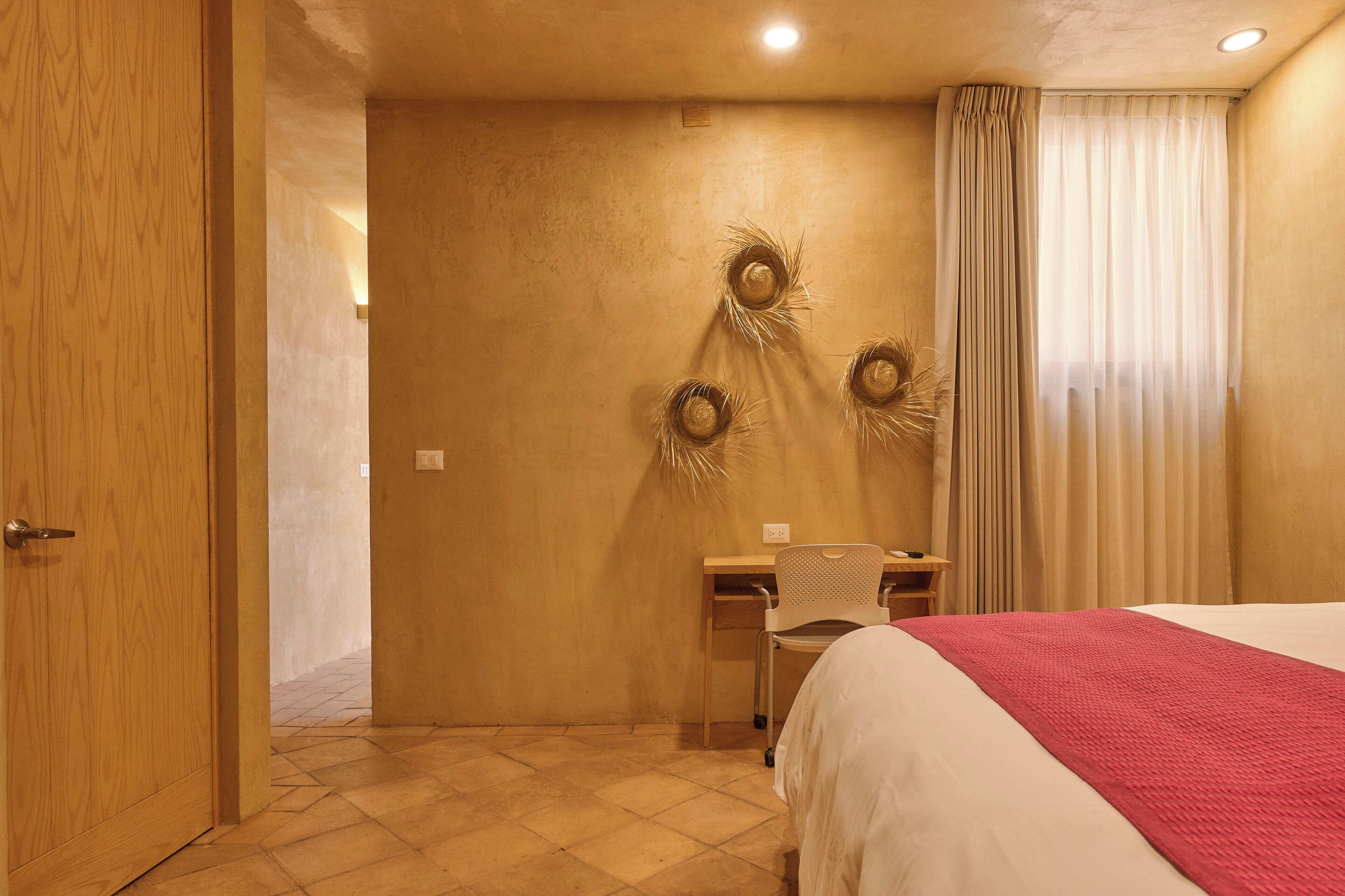 Ein Hotelzimmer mit Holztür, Schreibtisch mit Stuhl, Bett mit roter Decke und Wand mit drei Girlanden.