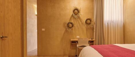 Ein Hotelzimmer mit Holztür, Schreibtisch mit Stuhl, Bett mit roter Decke und Wand mit drei Girlanden.