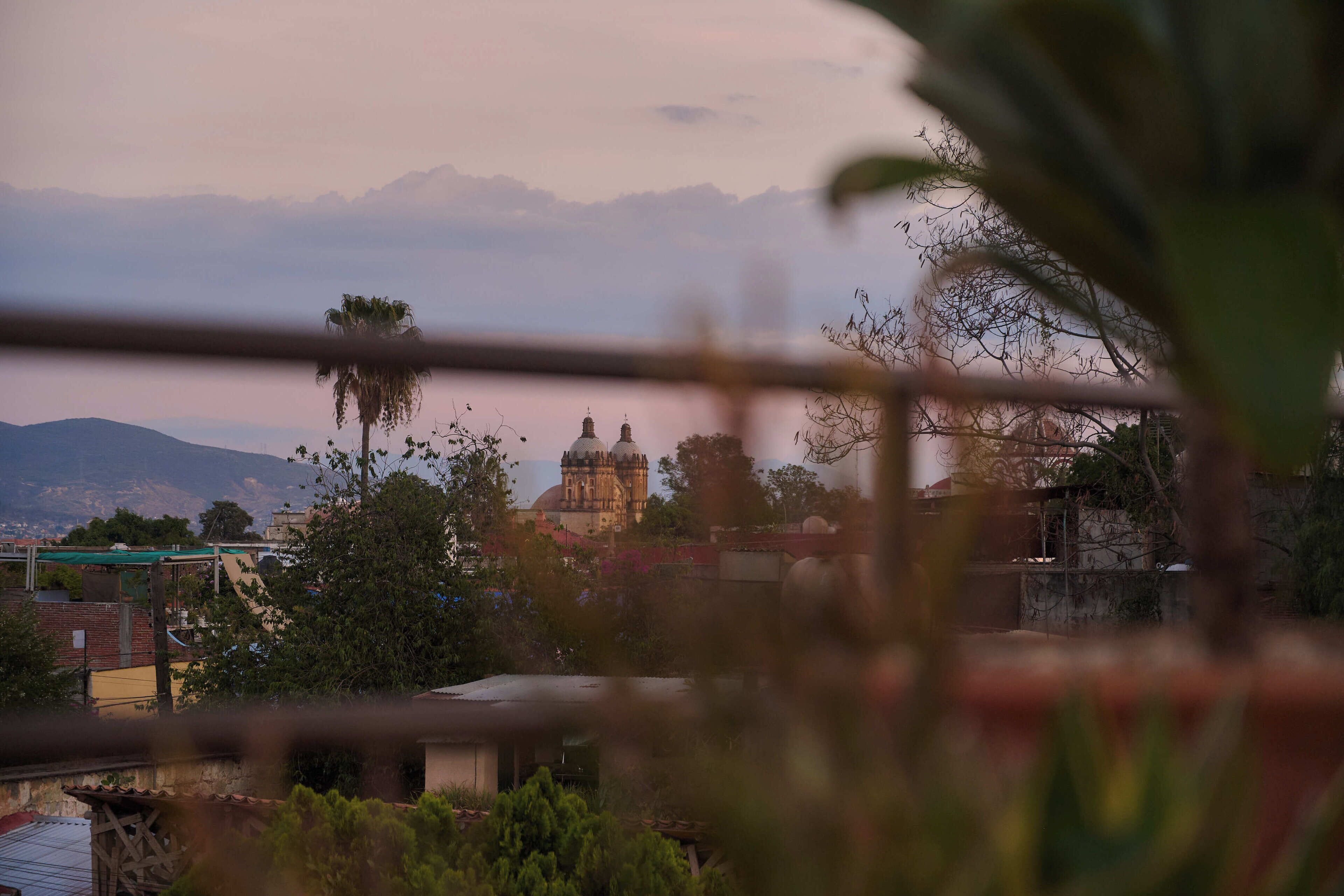 A view of a building with a dome and a tower, surrounded by greenery and mountains in the background.