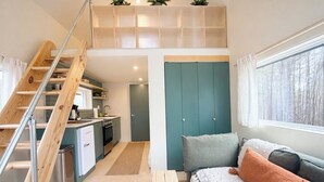 A compact living space with a kitchenette, a sofa with cushions, a staircase, and a view of trees through a window.