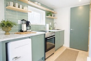 A compact kitchen with a built-in oven, a coffee maker, and a dishwasher. There are wooden shelves above the counter and a potted plant on the left.