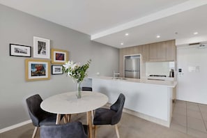 A modern kitchen with a central island, a dining area with a round table and chairs, and a wall with framed pictures. - Hamilton Portside Luxe – Level 5 (Brisbane)