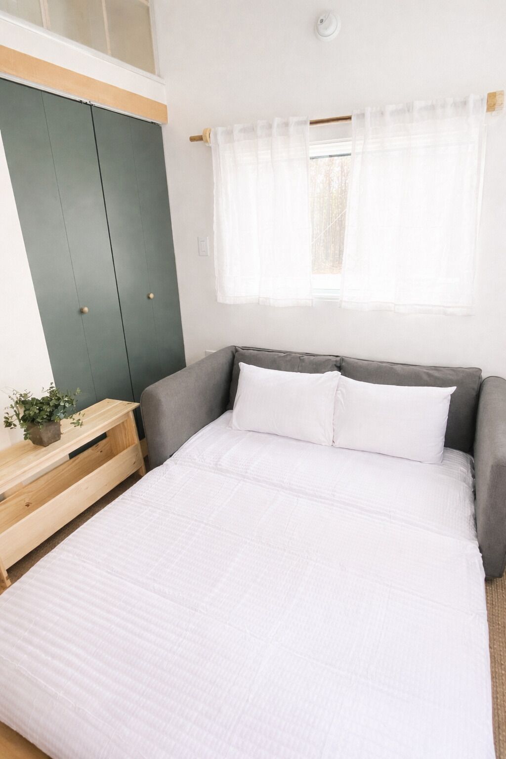 A bedroom with a bed, a bedside table with a plant, a window with curtains, and a wardrobe.