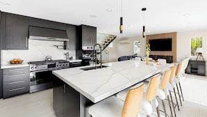 A modern kitchen with dark cabinetry, a large island, and a dining area with a high table and chairs. - Seal Beach Escape by Avantstay Modern Home + Fireplace Prvte Courtyard + Walk to Beach + Dining (Seal Beach)