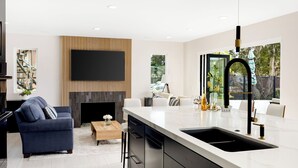 A modern kitchen with a large island, a flat-screen TV mounted on a wooden panel, and a dining area with white chairs. - Seal Beach Escape by Avantstay Modern Home + Fireplace Prvte Courtyard + Walk to Beach + Dining (Seal Beach)