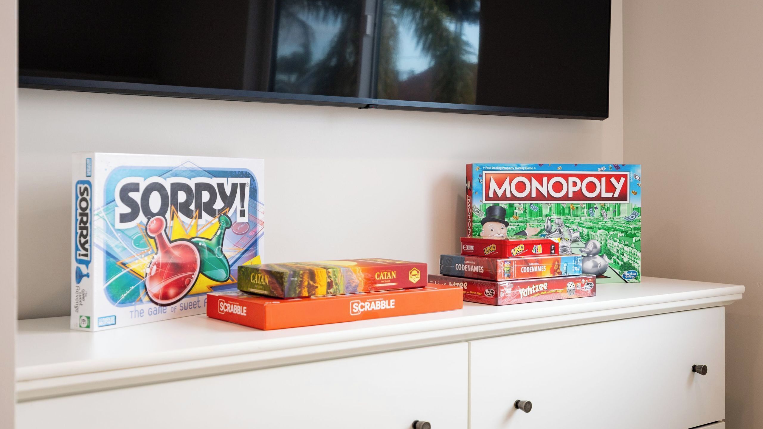 Un mueble blanco con un televisor montado encima. Sobre el mueble hay varios juegos de mesa, incluyendo 'Sorry!', 'Monopoly', 'Catan', 'Scrabble' y 'Uno'.