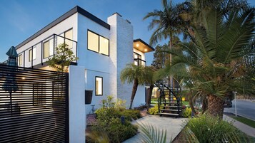Una casa moderna de dos pisos con un balcón, una escalera y un jardín bien cuidado con palmeras y arbustos.