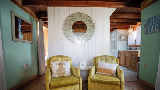 A room with two green armchairs, a wooden ceiling, a white wall with a decorative mirror, and a kitchen area in the background. - Pet-Friendly Coastal Cottage • Walk to Pier (Old Orchard Beach)