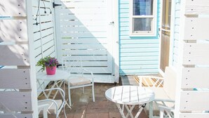 A small outdoor patio with white metal chairs and tables, a blue door, and hanging flower pots. - Pet-Friendly Coastal Cottage • Walk to Pier (Old Orchard Beach)