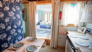 A kitchen with a stove, a dining table set with plates, and a view into the living room through a doorway. - Pet-Friendly Coastal Cottage • Walk to Pier (Old Orchard Beach)