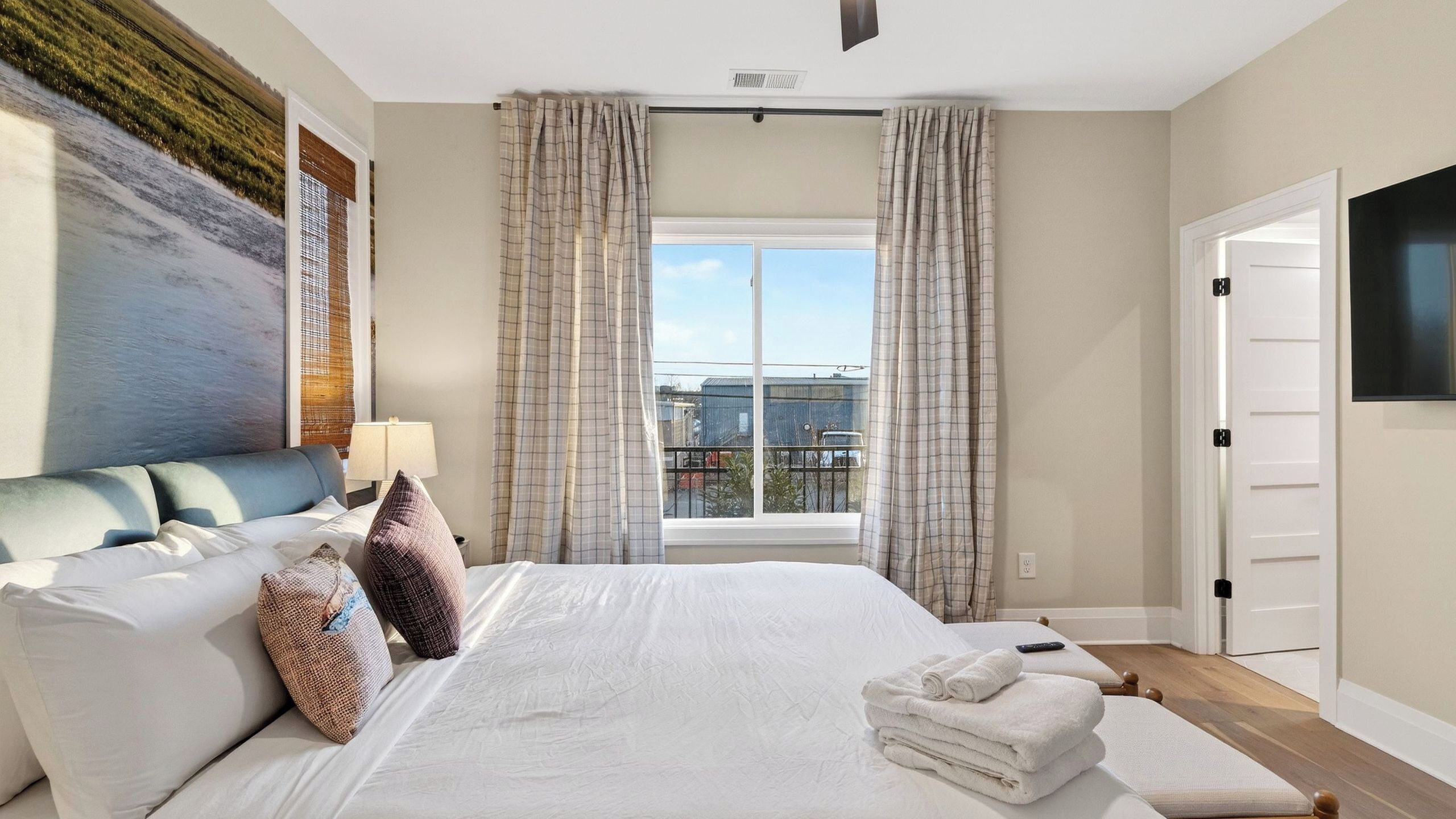 A hotel room with a large bed, a painting on the wall, a window with curtains, and a TV mounted on the wall.